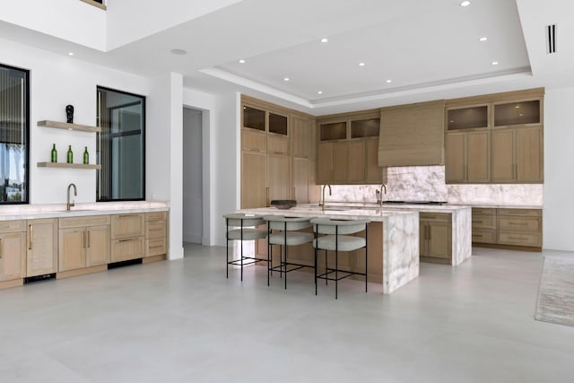 kitchen featuring a breakfast bar, a raised ceiling, a center island with sink, and wall chimney range hood