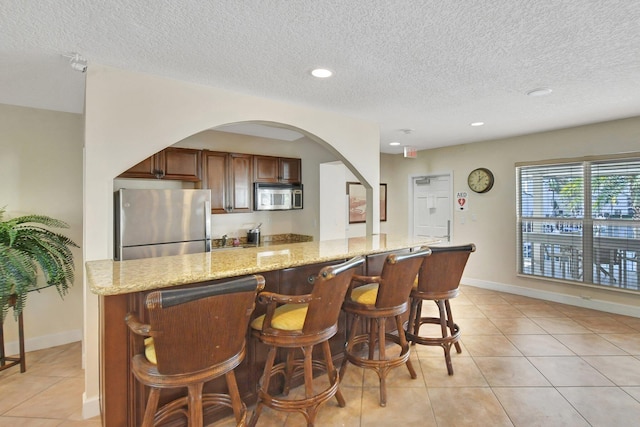 kitchen featuring stainless steel appliances, light stone counters, a textured ceiling, a breakfast bar area, and light tile patterned flooring