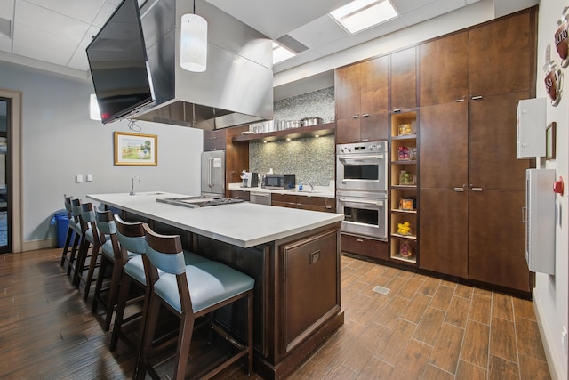 kitchen with a breakfast bar area, appliances with stainless steel finishes, dark hardwood / wood-style floors, island range hood, and a center island with sink