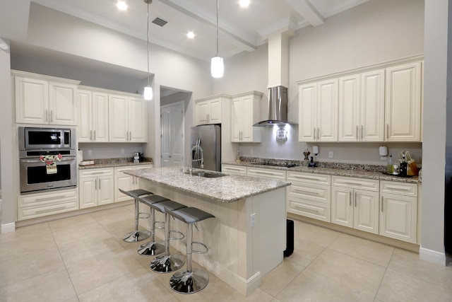 kitchen featuring a high ceiling, wall chimney range hood, an island with sink, appliances with stainless steel finishes, and light stone counters