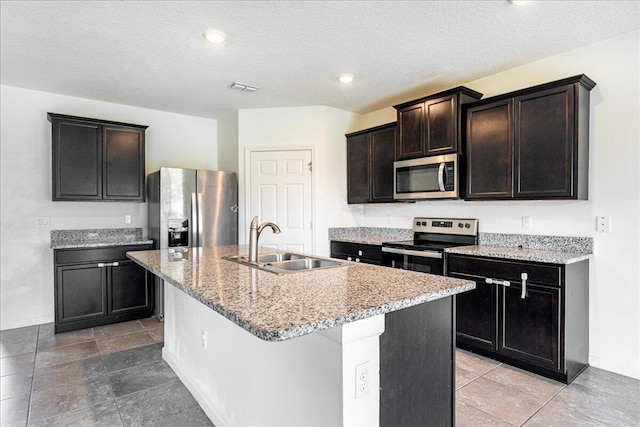 kitchen with appliances with stainless steel finishes, light stone counters, a textured ceiling, sink, and an island with sink