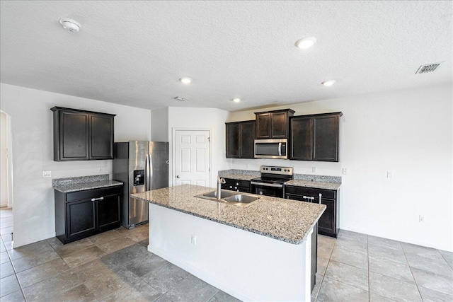 kitchen with light stone countertops, a textured ceiling, stainless steel appliances, sink, and an island with sink