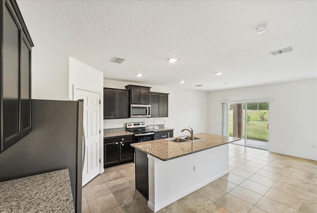 kitchen featuring sink, a textured ceiling, an island with sink, appliances with stainless steel finishes, and light stone counters