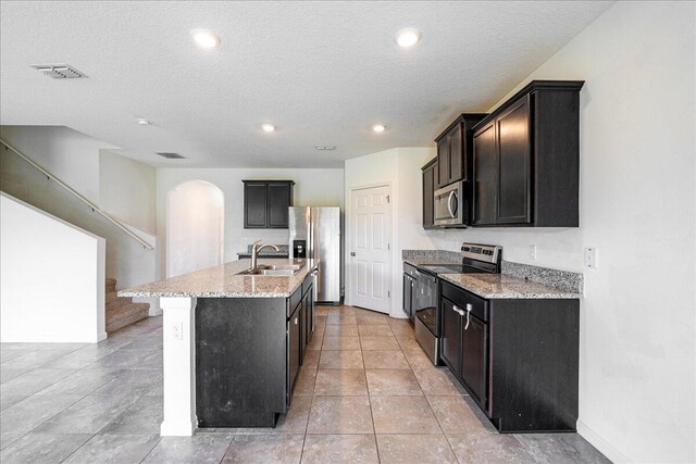 kitchen with a center island with sink, sink, a textured ceiling, appliances with stainless steel finishes, and light stone counters