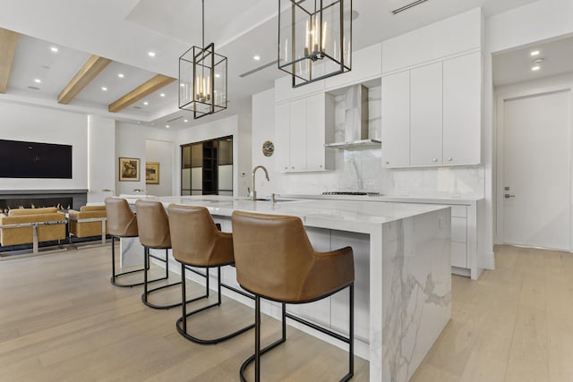 kitchen with backsplash, wall chimney exhaust hood, a large island with sink, beamed ceiling, and hanging light fixtures