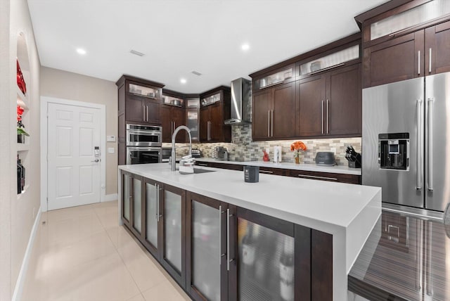 kitchen with stainless steel appliances, wall chimney range hood, an island with sink, decorative backsplash, and dark brown cabinets