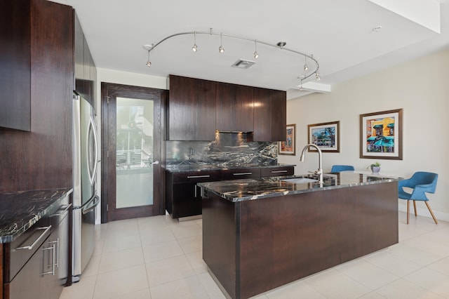 kitchen with stainless steel refrigerator, dark stone countertops, a kitchen island with sink, and decorative backsplash