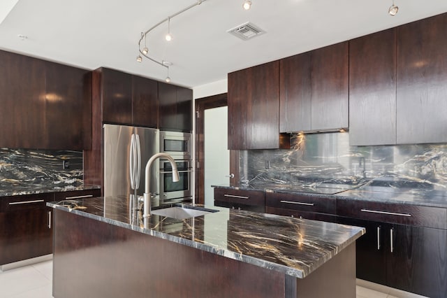 kitchen featuring sink, stainless steel appliances, backsplash, dark stone countertops, and a center island with sink