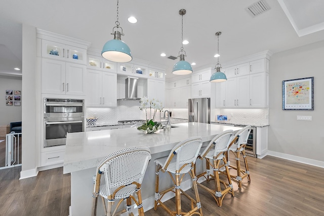 kitchen with appliances with stainless steel finishes, wall chimney exhaust hood, decorative light fixtures, white cabinetry, and an island with sink