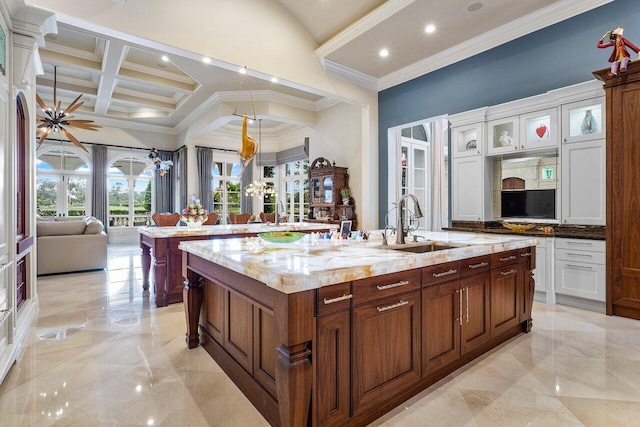 kitchen featuring light stone countertops, coffered ceiling, ornamental molding, and an island with sink