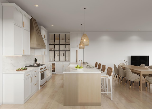 kitchen with custom range hood, light wood-type flooring, hanging light fixtures, and an island with sink