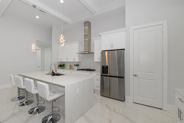 kitchen featuring wall chimney range hood, sink, a kitchen island with sink, hanging light fixtures, and stainless steel fridge with ice dispenser