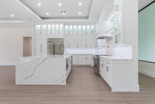 kitchen featuring premium range hood, a raised ceiling, light stone countertops, premium appliances, and white cabinetry