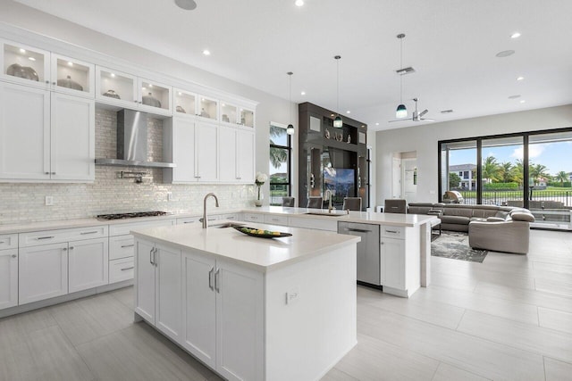 kitchen with a center island with sink, white cabinets, wall chimney range hood, hanging light fixtures, and appliances with stainless steel finishes