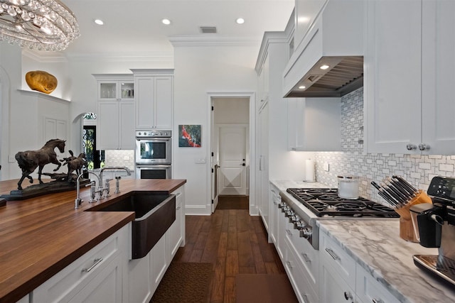 kitchen with white cabinetry, butcher block countertops, decorative backsplash, custom exhaust hood, and appliances with stainless steel finishes