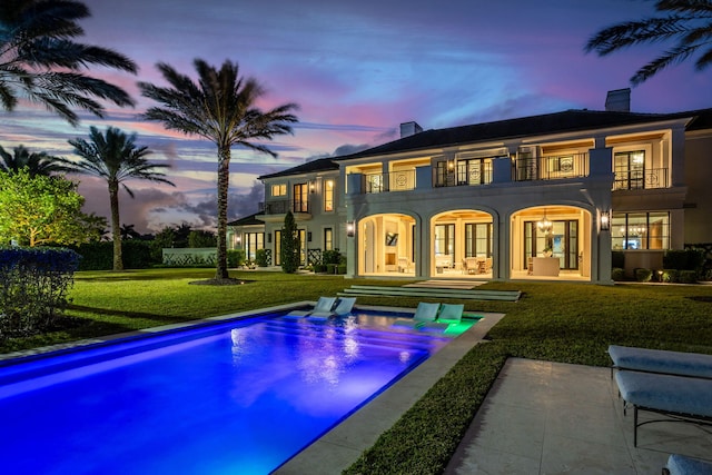 back house at dusk featuring a patio, a balcony, a pool with hot tub, and a lawn