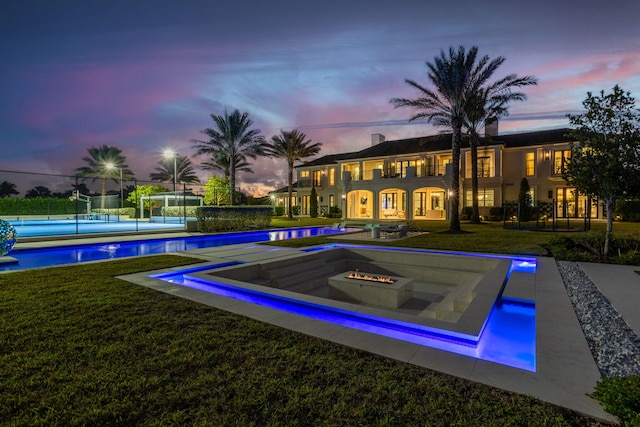 pool at dusk featuring an outdoor fire pit, a yard, and tennis court