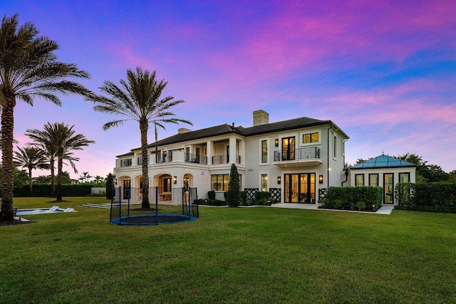 back house at dusk with a trampoline, a lawn, french doors, and a balcony