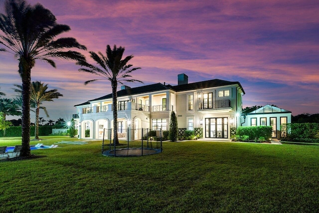 back house at dusk featuring french doors, a balcony, a trampoline, and a lawn