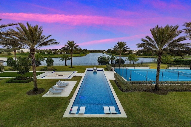 pool at dusk with tennis court, a lawn, and a water view