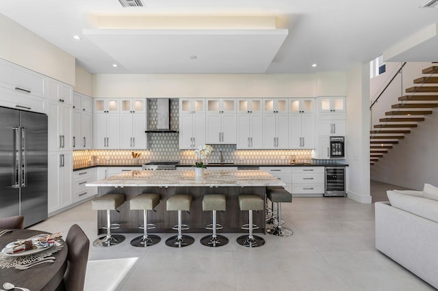 kitchen with a center island, wall chimney exhaust hood, high end refrigerator, a kitchen bar, and white cabinets