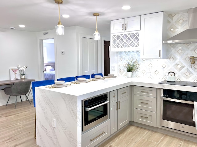 kitchen featuring light wood finished floors, a peninsula, oven, decorative backsplash, and wall chimney exhaust hood