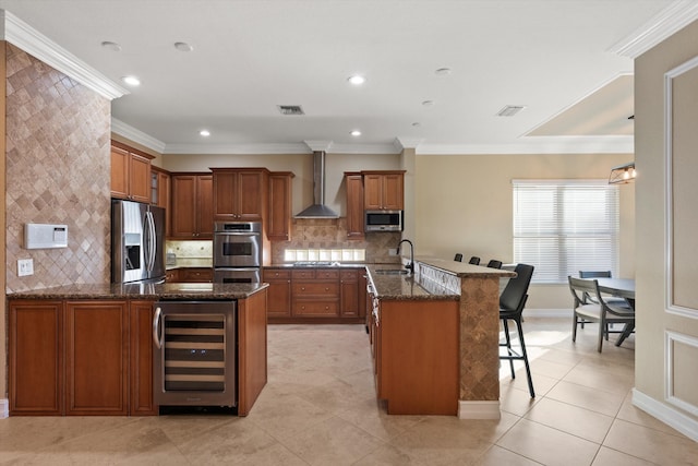 kitchen with wall chimney range hood, stainless steel appliances, an island with sink, a kitchen bar, and beverage cooler