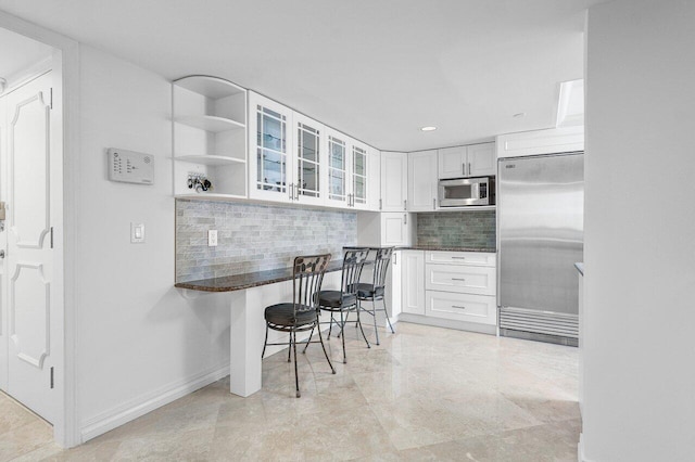 kitchen featuring built in appliances, decorative backsplash, white cabinets, dark stone countertops, and a kitchen breakfast bar