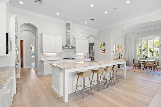 kitchen with a kitchen island with sink, wall chimney range hood, ornamental molding, white cabinetry, and gas cooktop