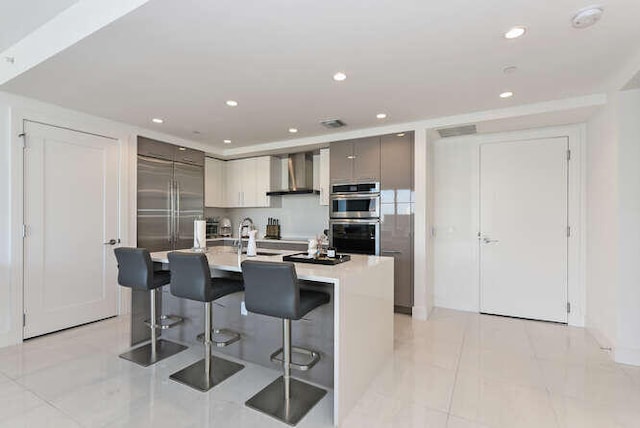 kitchen featuring gray cabinetry, a breakfast bar, a kitchen island with sink, wall chimney exhaust hood, and stainless steel appliances