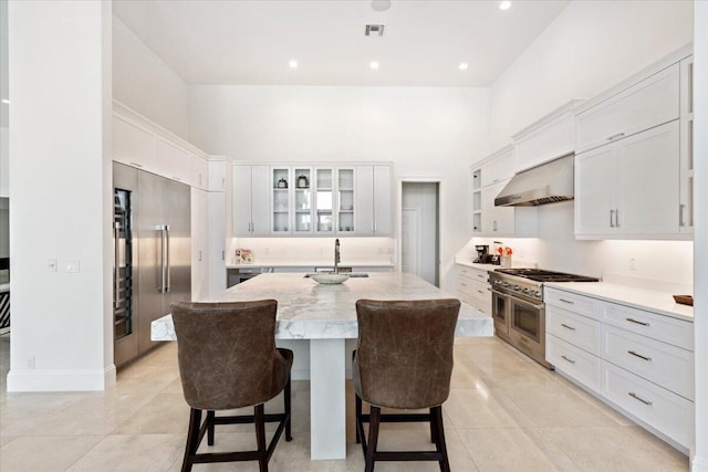 kitchen featuring a kitchen island with sink, wall chimney exhaust hood, premium appliances, light stone counters, and a breakfast bar area