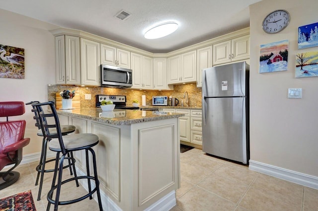 kitchen featuring stainless steel appliances, light stone counters, kitchen peninsula, cream cabinetry, and a breakfast bar