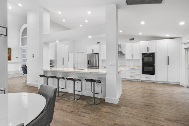 kitchen featuring wall chimney exhaust hood, black double oven, high vaulted ceiling, white cabinets, and stainless steel fridge with ice dispenser