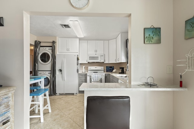 kitchen featuring white appliances, kitchen peninsula, stacked washer / drying machine, light tile patterned flooring, and white cabinetry