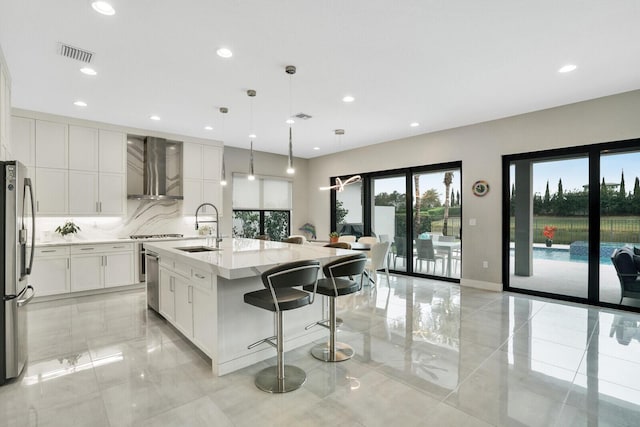 kitchen with stainless steel refrigerator with ice dispenser, hanging light fixtures, a kitchen island with sink, wall chimney exhaust hood, and light stone counters