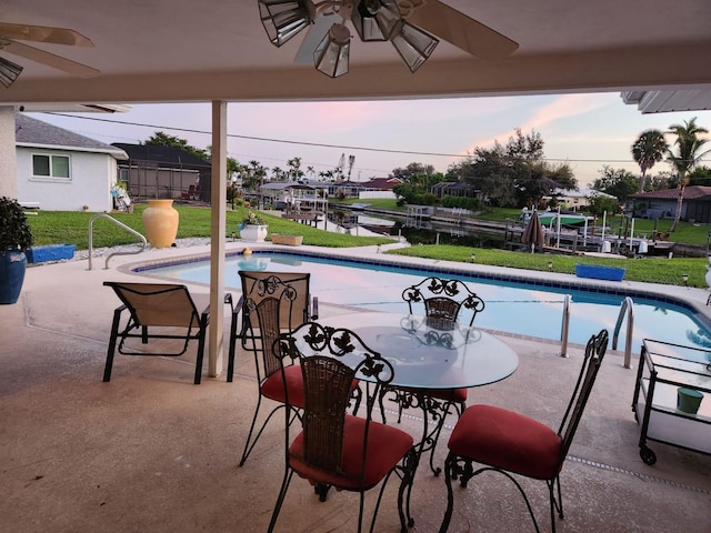 pool at dusk featuring a lawn, ceiling fan, and a patio area