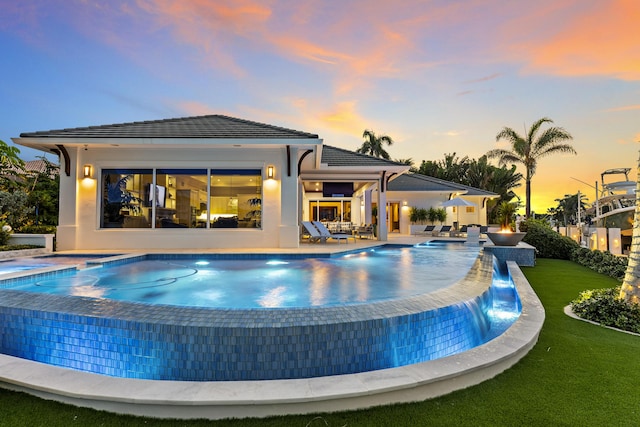 pool at dusk with a lawn, pool water feature, and a hot tub