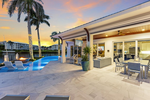 pool at dusk with a fire pit, ceiling fan, a patio area, and a water view