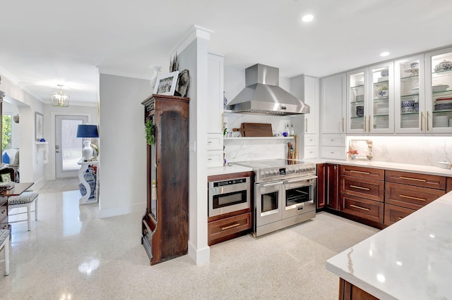 kitchen featuring light stone counters, ornamental molding, wall chimney range hood, range with two ovens, and white cabinetry