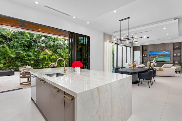 kitchen with light stone counters, hanging light fixtures, a center island with sink, a notable chandelier, and sink