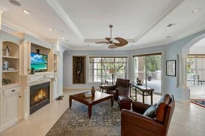 living room with built in shelves, ceiling fan, a raised ceiling, light tile patterned floors, and ornamental molding