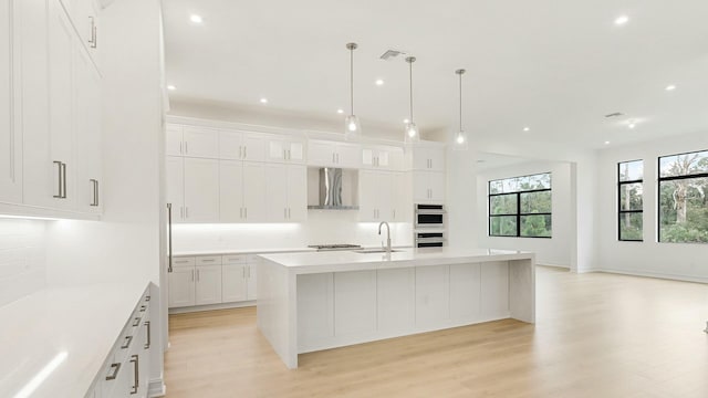 kitchen with wall chimney range hood, sink, decorative light fixtures, and a center island with sink