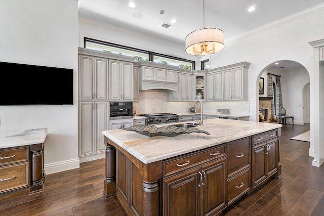 kitchen with dark wood-type flooring, a stone fireplace, decorative backsplash, light stone countertops, and an island with sink