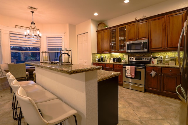 kitchen featuring a kitchen bar, appliances with stainless steel finishes, a kitchen island with sink, decorative light fixtures, and dark stone countertops