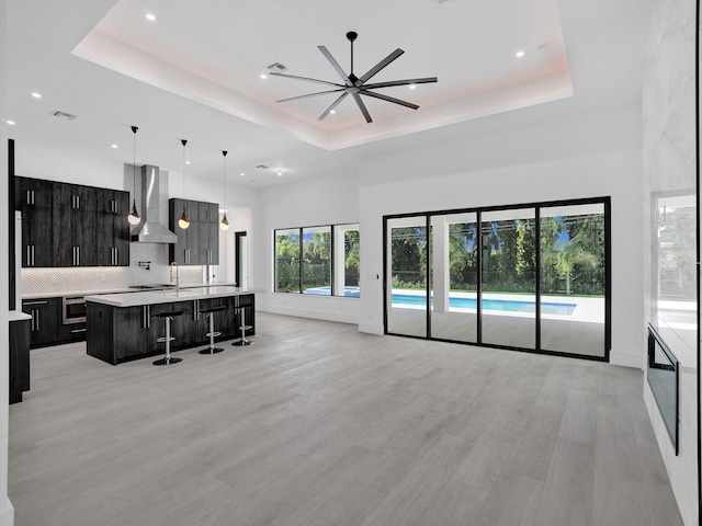 kitchen featuring a kitchen island with sink, a kitchen breakfast bar, a raised ceiling, wall chimney range hood, and ceiling fan