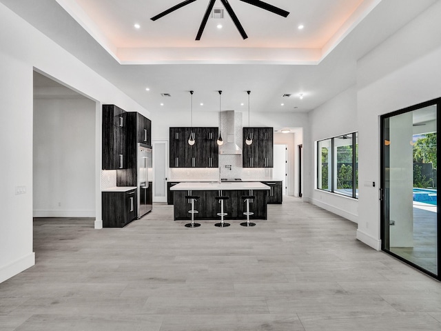 kitchen with ceiling fan, wall chimney range hood, pendant lighting, a center island with sink, and a breakfast bar area