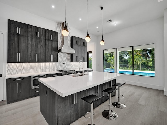 kitchen featuring a kitchen bar, wall oven, wall chimney exhaust hood, hanging light fixtures, and an island with sink