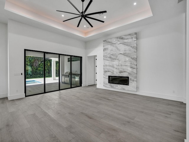 unfurnished living room featuring ceiling fan, a premium fireplace, a towering ceiling, a tray ceiling, and light hardwood / wood-style floors