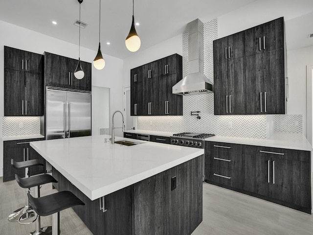 kitchen with sink, wall chimney exhaust hood, hanging light fixtures, stainless steel appliances, and a kitchen island with sink