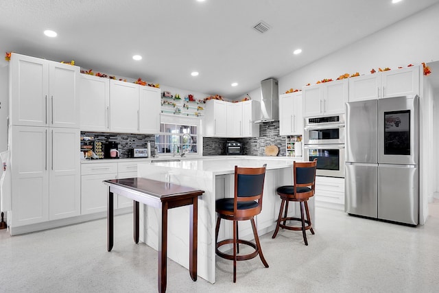 kitchen with a center island, white cabinets, a kitchen breakfast bar, wall chimney range hood, and appliances with stainless steel finishes